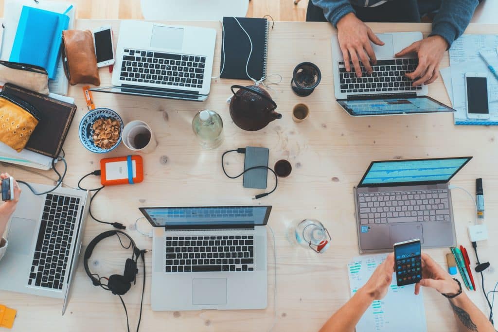 Des ordinateurs et du matériel de bureautique sont posés sur une table. Des collaborateurs travaillent dessus, seules leurs mains dépassent. 