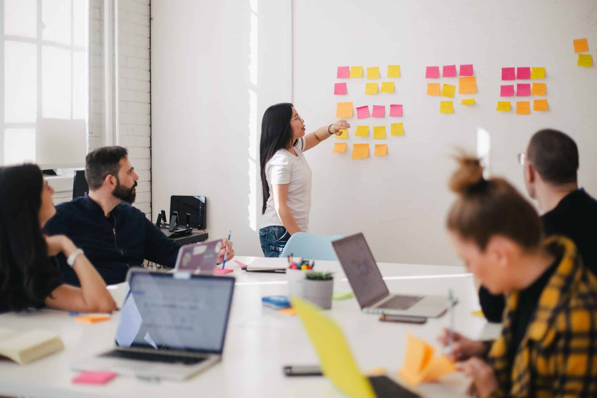 Une consultante anime une réunion, en désignant des post-it collés sur un tableau blanc. Les collaborateurs de l'entreprise concernée l'écoutent attentivement. 