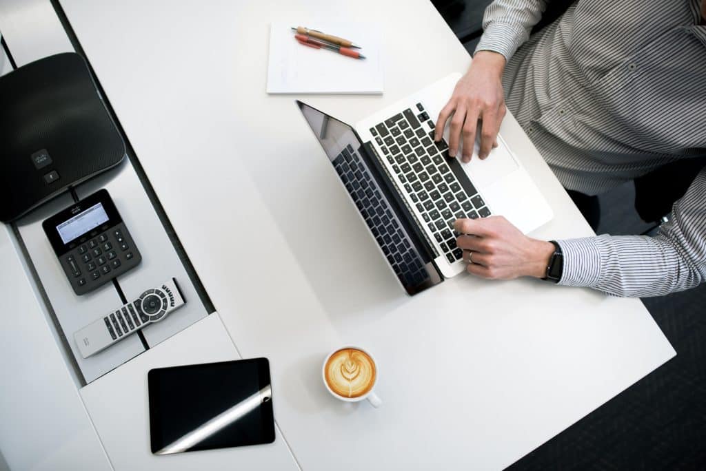 Un homme tape sur le clavier de son ordinateur, à son bureau. Un café, une tablette tactile et deux stylos sont posés sur la table.