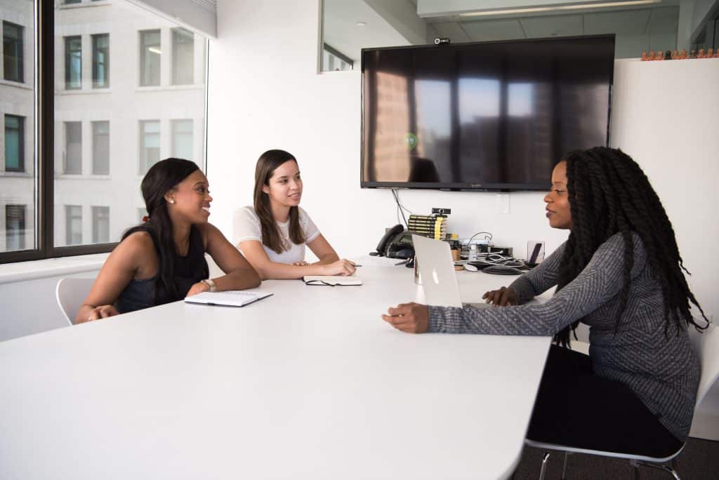 Une équipe de RH discute autour d'une table de travail, avec un ordinateur et une télé éteinte en fonds.